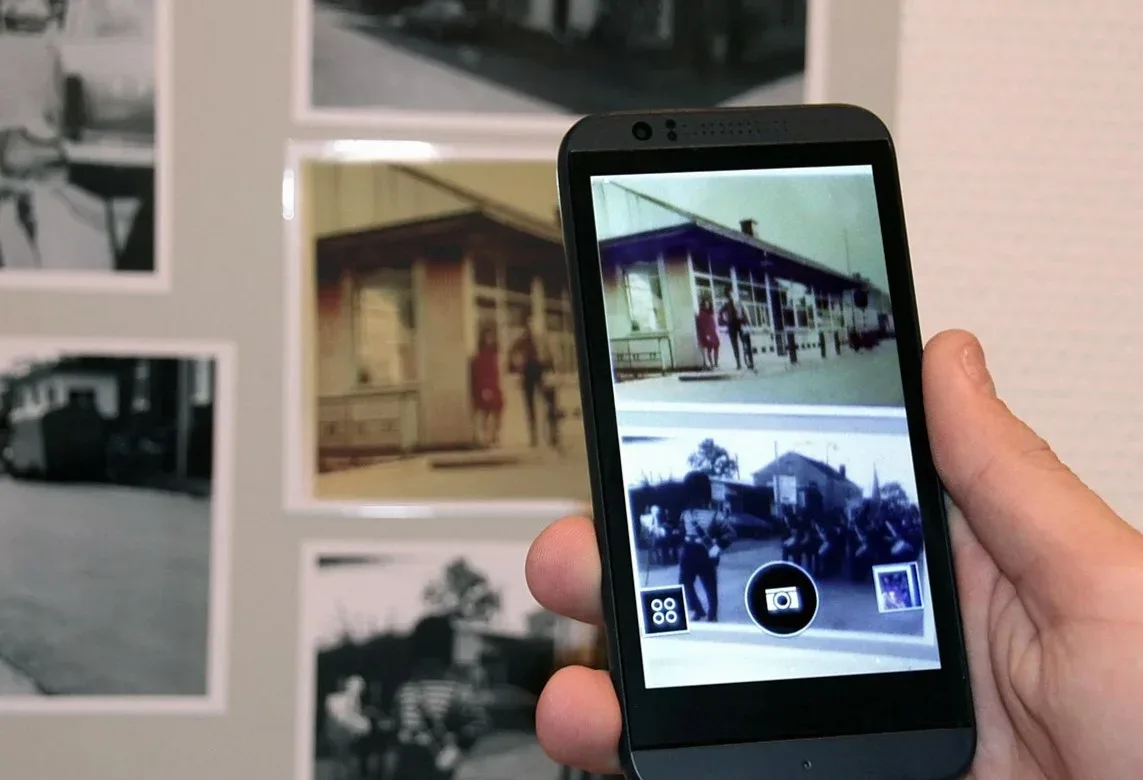 スマートフォンの画面に古い写真を映した手元と、壁に並ぶ写真